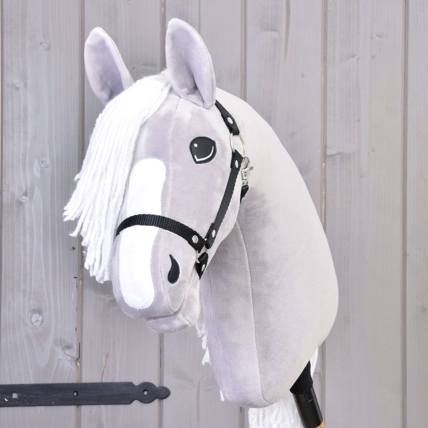 Hobby Horse LarDen Hobby Horse Popcorn mit Schwarzem Halfter mit grauem Plüschdesign, weißer Mähne und freundlichem Gesicht – ideal als Begleiter für fantasievolles Spiel. Hier vor einer hellen Holzwand gezeigt.