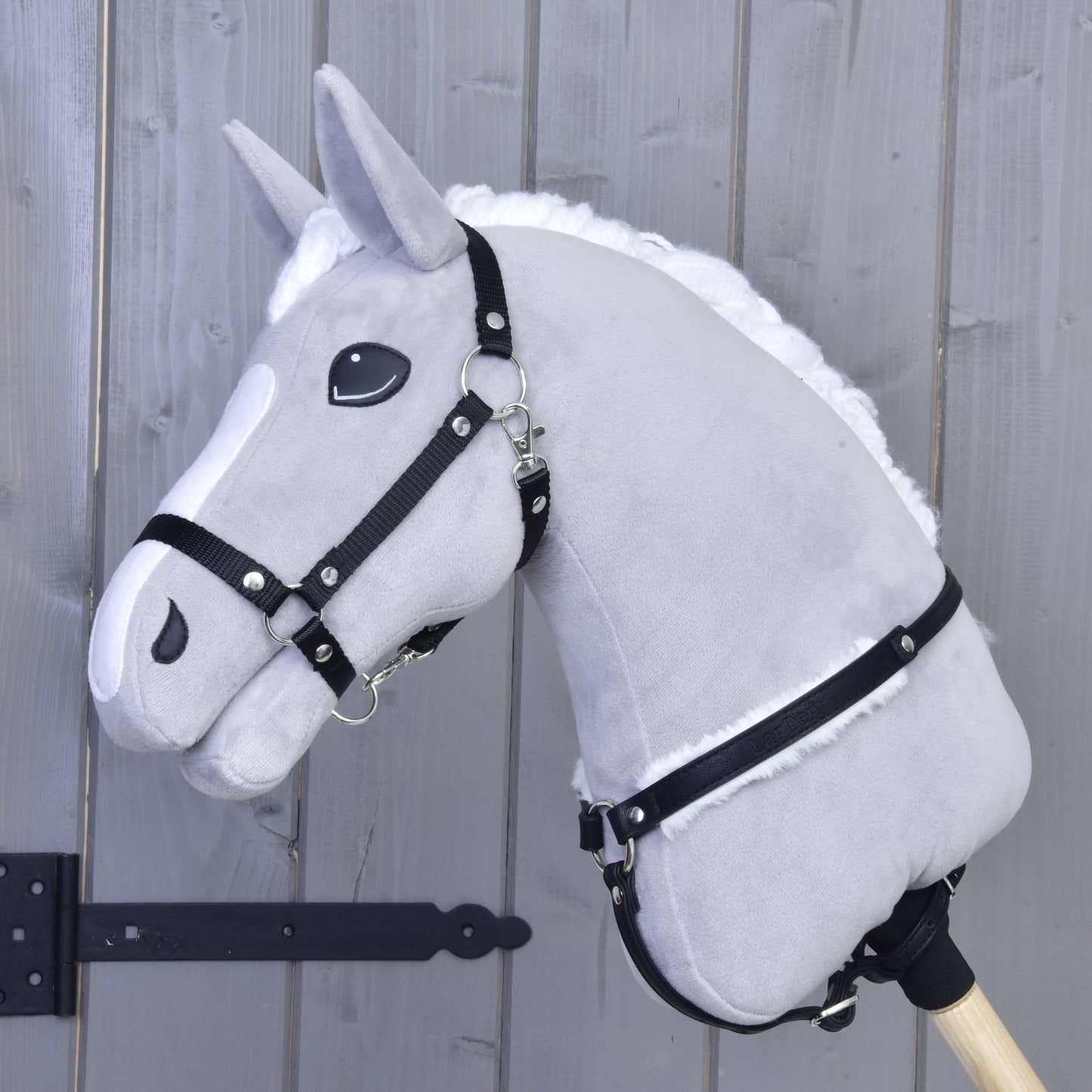 The Hobby Horse LarDen Breastgirth with fur Espresso is a plush gray hobby horse with a white mane, black bridle, and soft fur-lined breastgirth on a wooden stick, shown against a light wood-paneled wall.