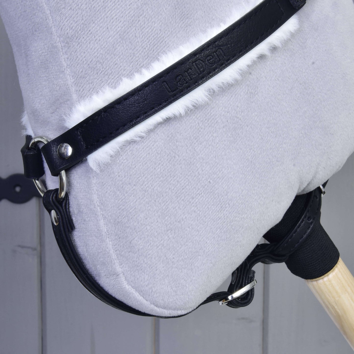 Close-up of a Hobby Horse LarDen toy with a soft light gray head, black faux leather bridle, and the "Breastgirth with fur Espresso" accessory, set against a wooden background.