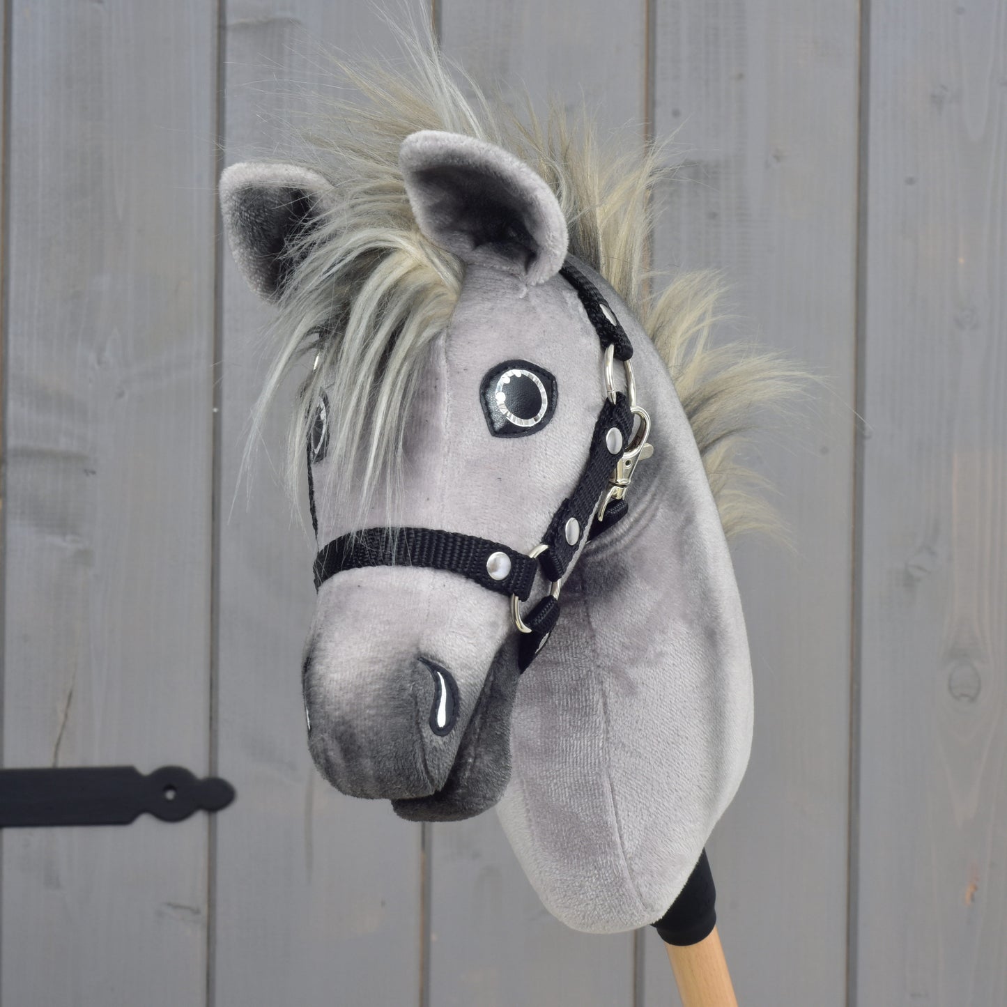 A Hobby Horse LarDen Hobby Horse Prestige with black halter, size XS, featuring a gray plush body with a blond mane and mounted on a wooden stick, is displayed in front of a light brown wooden wall.