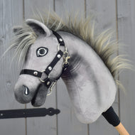 Hobby Horse Prestige mit schwarzem Halfter, Größe XS von Hobby Horse LarDen mit grauem Plüschdesign, struppiger Mähne und schwarzem Halfter, inspiriert von der Eleganz eines englischen Warmbluts. Es ist auf einem Holzstab befestigt und vor einem Hintergrund aus Holzpaneelen dargestellt.