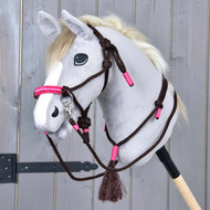 Seilset Noir Cocoa von Hobby Horse LarDen mit plüschigem Pferdekopf mit weißem und grauem Fell, schwarzen Knopfaugen, cremefarbener Mähne sowie braun-pinkem Halfter für Hobby Horse mit Zügeln, auf einem Holzstab vor Paneelhintergrund.