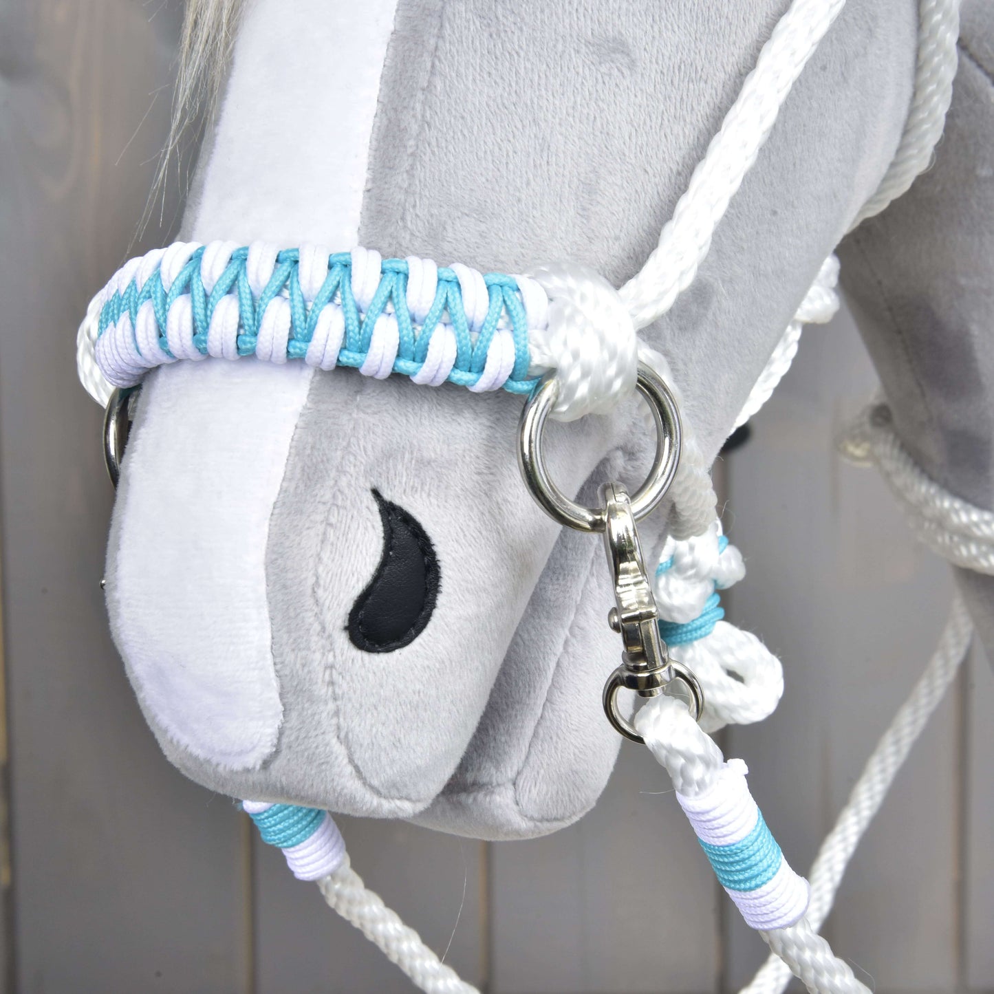 Gros plan sur une tête en peluche grise Hobby Horse LarDen avec l’ensemble de corde Reine Blanche, un licol et des rênes tressés blancs, bleus et violet clair, avec œil noir brodé et anneau métallique sur le harnachement.