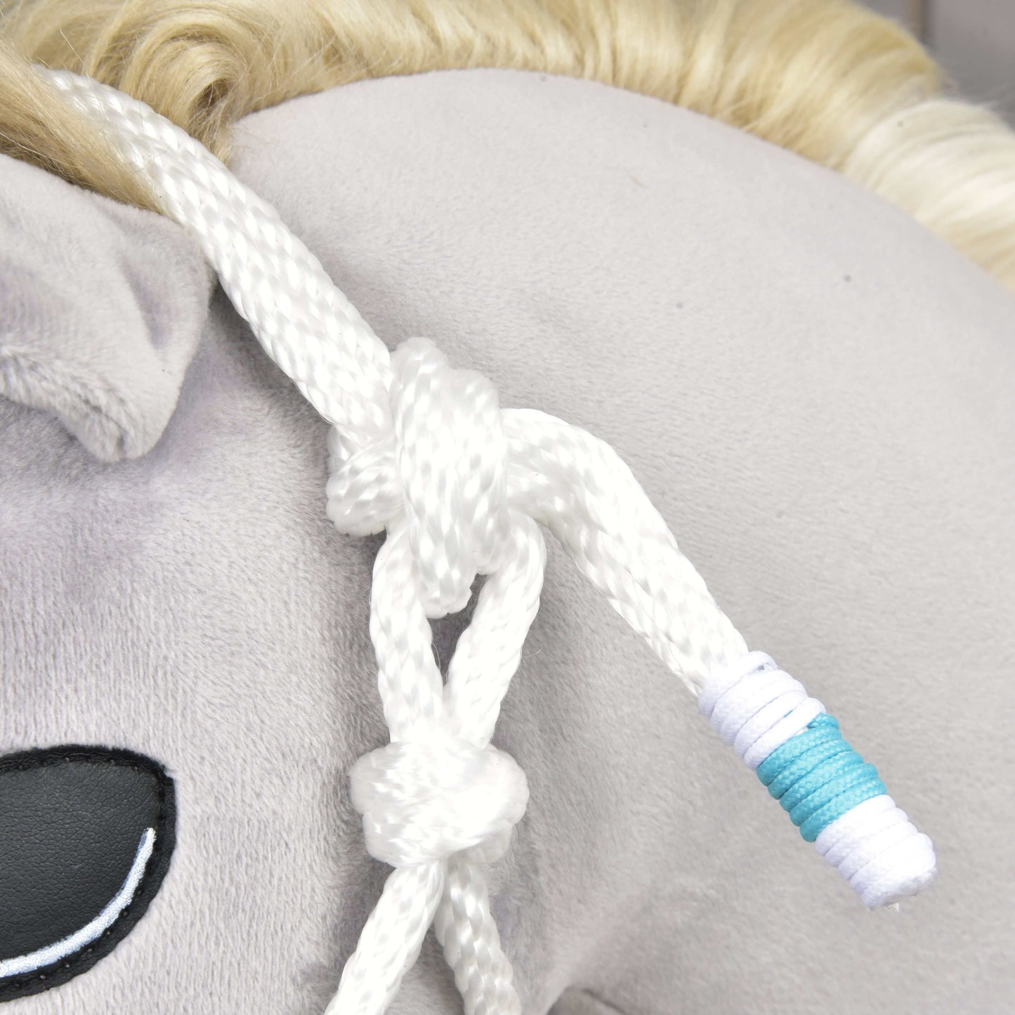 Gros plan sur un Hobby Horse LarDen en tissu gris clair, avec œil noir brodé, crinière blond clair et l’ensemble de corde Reine Blanche, un licol en corde blanche noué et enroulé de fil bleu et blanc.