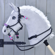 Seilset Princess von Hobby Horse LarDen mit grauem Hobby Horse mit weißer Mähne, detailliertem schwarzem Halfter und Zügeln sowie pinken und türkisen Akzenten, auf einem Holzstab vor grauem Plankenhintergrund.