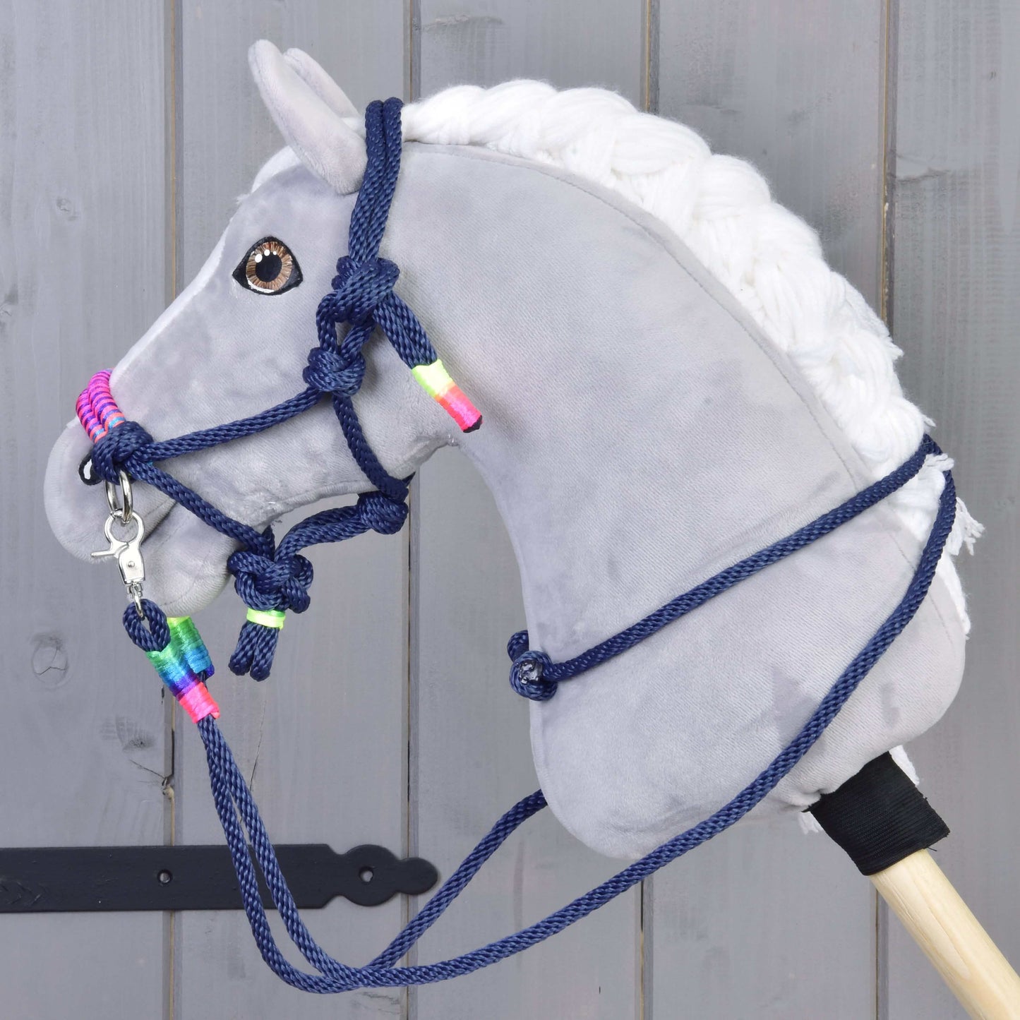 Seilset Rosemary von Hobby Horse LarDen mit grauem Plüsch-Hobby Horse mit weißer Mähne, dunkelblauen Zügeln und Halfter mit Neonperlen, montiert auf einem Holzstab vor grauem Holzhintergrund.