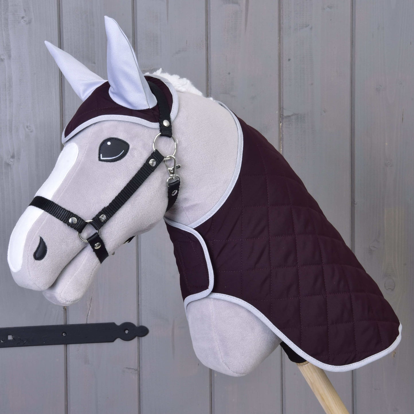 Hobby Horse LarDen Set Purple mit grauem Hobby Horse, gesteppter dunkelvioletter Decke, Zaumzeug und Fliegenhaube, alles auf einem Holzstab vor einem Hintergrund aus Holzplanken.