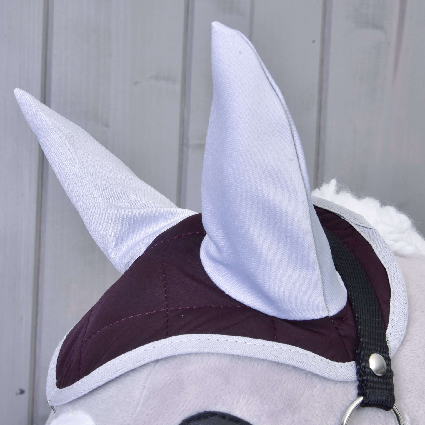 Nahaufnahme von Hobby Horse LarDen Set Purple: ein plüschiger Pferdekopf mit großen grauen Ohren, kastanienrotem und grauem gestepptem Zaumzeug und Fliegenhaube. Im Hintergrund vertikale hellgraue Holzpaneele.