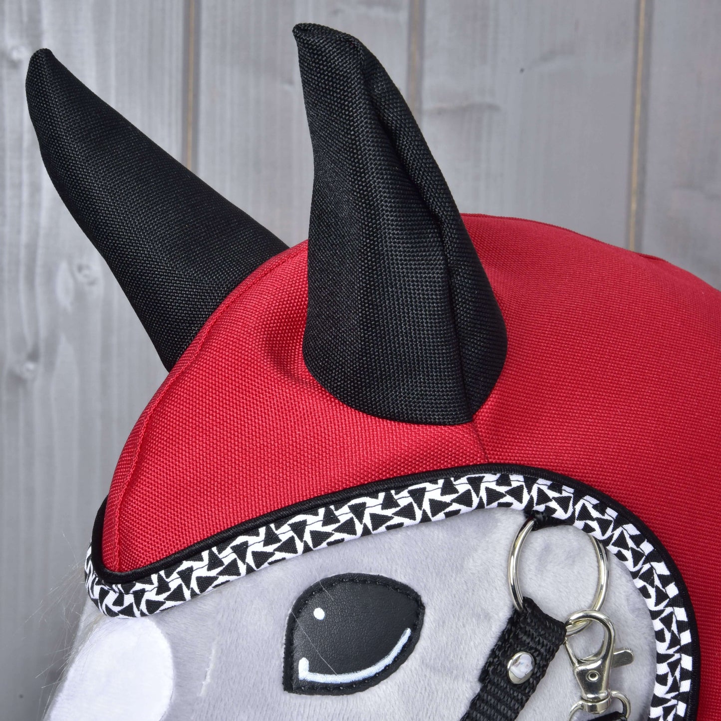 Nahaufnahme des Hobby Horse LarDen „Regendecke Red Ferrari“ Plüschspielzeugs mit roter und schwarzer Kapuze mit spitzen Ohren, dekorativem Besatz, Zaumzeug und gestickten Augen, einer Allwetter-Regendecke ähnlich, vor Holzhintergrund.