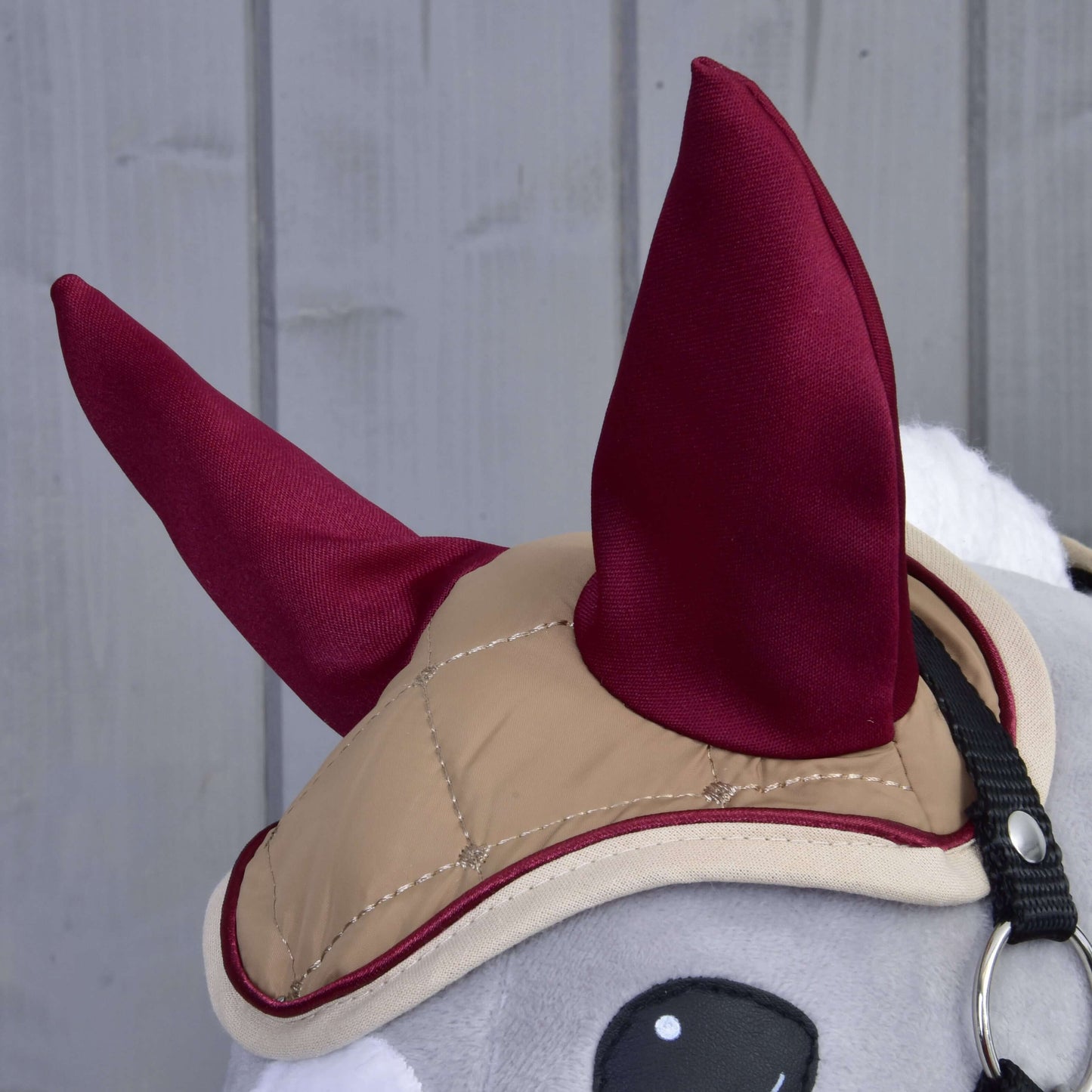 Nahaufnahme des grauen Set Roma Plüschspielzeugs von Hobby Horse LarDen mit hellbraunem Zaumzeug und bordeauxfarbener Fliegenhaube vor grauem Holzhintergrund.