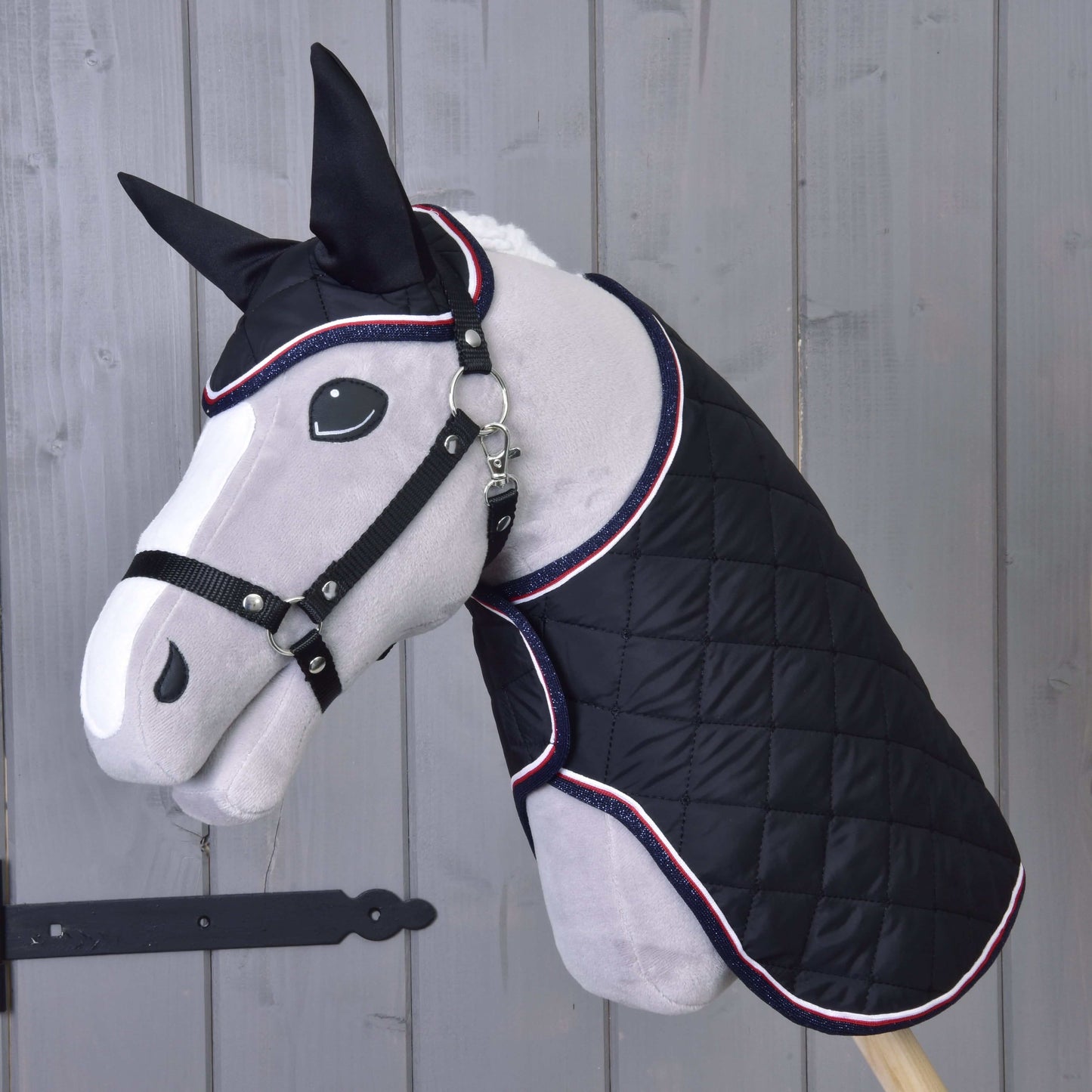 Ein graues Plüschpferd-Modell präsentiert das Hobby Horse LarDen Set Royal, eine schwarze gesteppte Decke und passende Fliegenhaube. Im Hintergrund eine helle Holzwand mit schwarzem Metallriegel links.