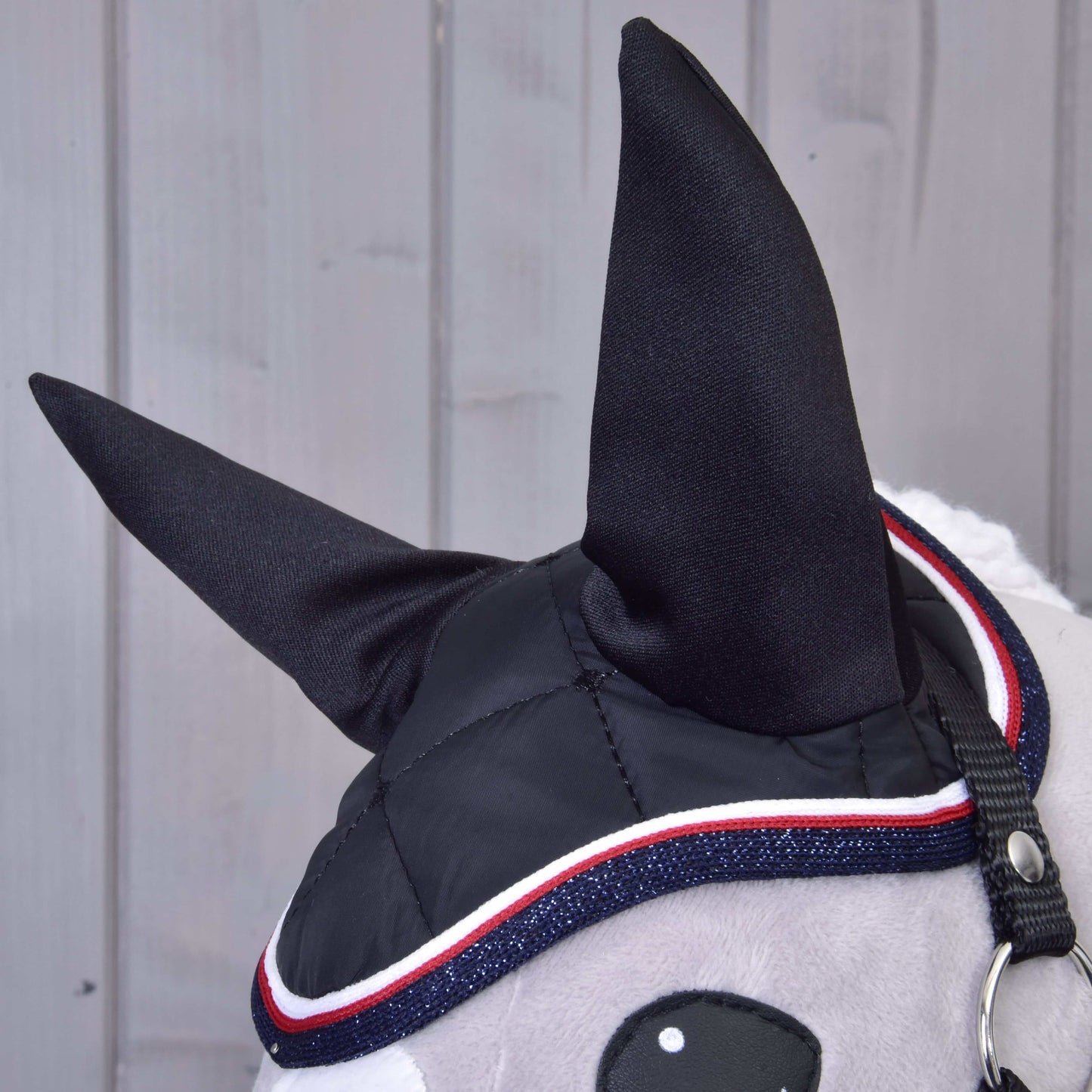 Nahaufnahme von Hobby Horse LarDen Set Royal: ein Hobby Horse Kopf mit schwarzen Stoffohren, stilvoller Fliegenhaube mit rotem, weißem und blauem Besatz, grauem Gesicht und schwarzem Zaumzeug vor hellem Holz-Hintergrund.