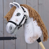 Hobby Horse LarDen „Hobby Horse Rufus mit Schwarzem Halfter“ mit handgefertigtem gesprenkeltem weißem Stoffkopf, brauner Garnmähne, Knopfaugen und schwarzem Halfter für Hobby Horse auf einem Holzstab vor grauem Holz-Hintergrund.