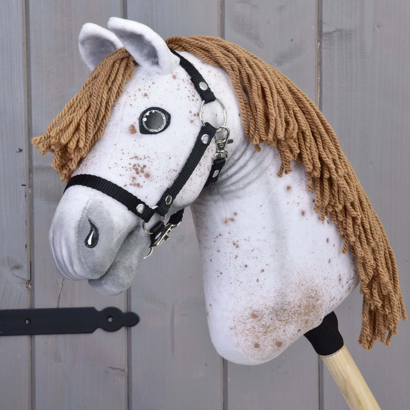 Hobby Horse LarDen „Hobby Horse Rufus mit Schwarzem Halfter“ mit handgefertigtem gesprenkeltem weißem Stoffkopf, brauner Garnmähne, Knopfaugen und schwarzem Halfter für Hobby Horse auf einem Holzstab vor grauem Holz-Hintergrund.