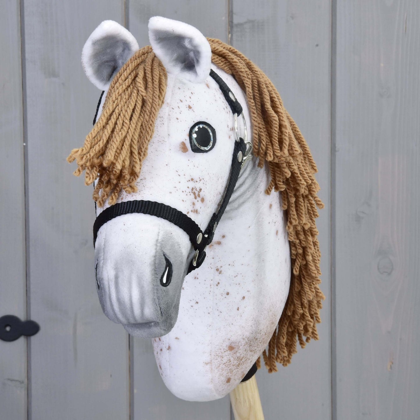 Hobby Horse Rufus avec licol noir de Hobby Horse LarDen avec visage blanc en peluche fait main, crinière brune en fil, licol noir, détails peints, monté sur un bâton en bois devant un fond de panneaux en bois gris.