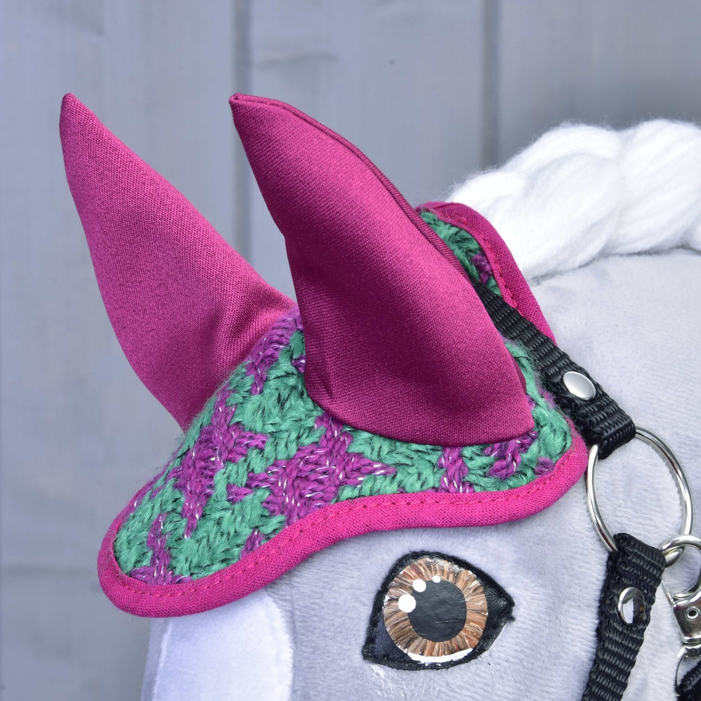 Eine Nahaufnahme von Hobby Horse LarDen Set Coco zeigt den grauen Kopf mit einer gehäkelten Fliegenhaube in Grün und Lila, großen rosa Stoffohren, weißer Mähne und schwarzem Zaumzeug mit glänzendem Metallring.