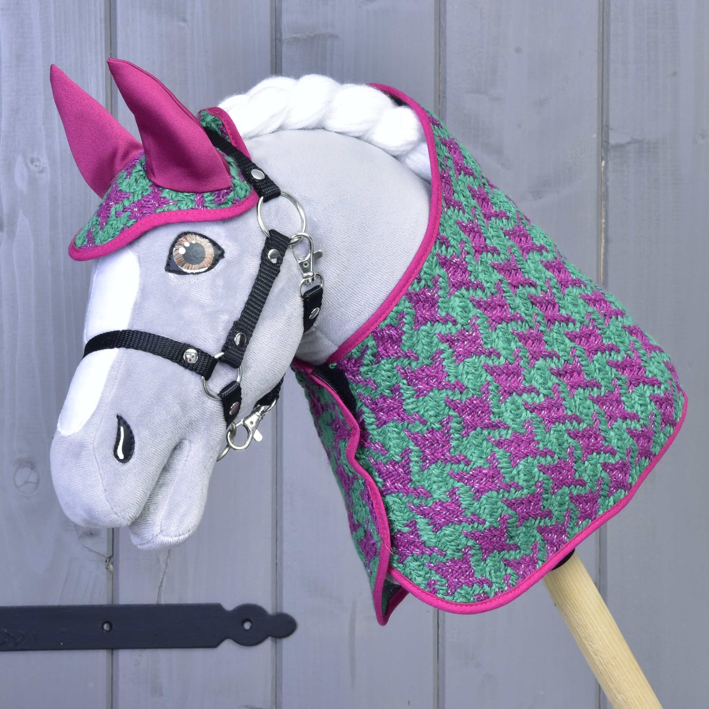 Ein graues Hobby Horse LarDen steht vor einer hellgrauen Holzwand und trägt das Set Coco: ein Deckenset mit Hahnentrittmuster in Rosa und Grün mit passender rosa Fliegenhaube und schwarzem Zaumzeug.