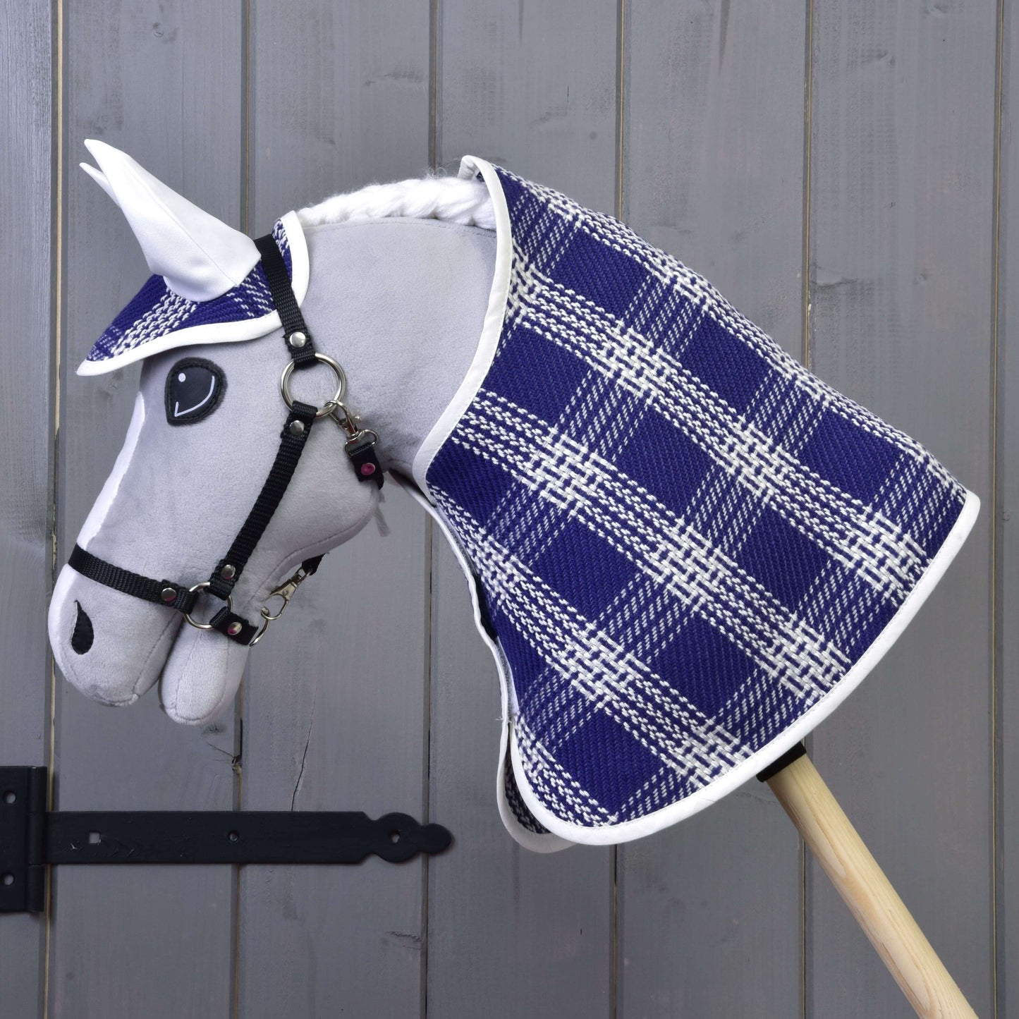 Das Hobby Horse LarDen "Set Blue Cube" zeigt einen grauen Plüschkopf, ein blau-weiß kariertes Deckenset mit passender Fliegenhaube, montiert auf einem Holzstab vor einer grauen Holzwand.