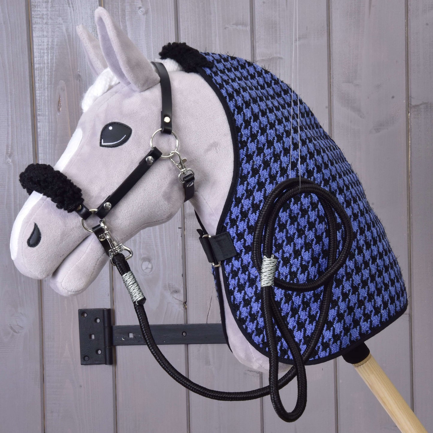 Ein Hobby Horse LarDen Set Harmony Blue Plüsch-Hobby Horse mit schwarzem Halfter für Hobby Horse, Zügeln und einer blau-schwarzen Stalldecke mit Hahnentrittmuster ist an einer Wand vor einem Hintergrund aus hellen Holzplanken montiert.