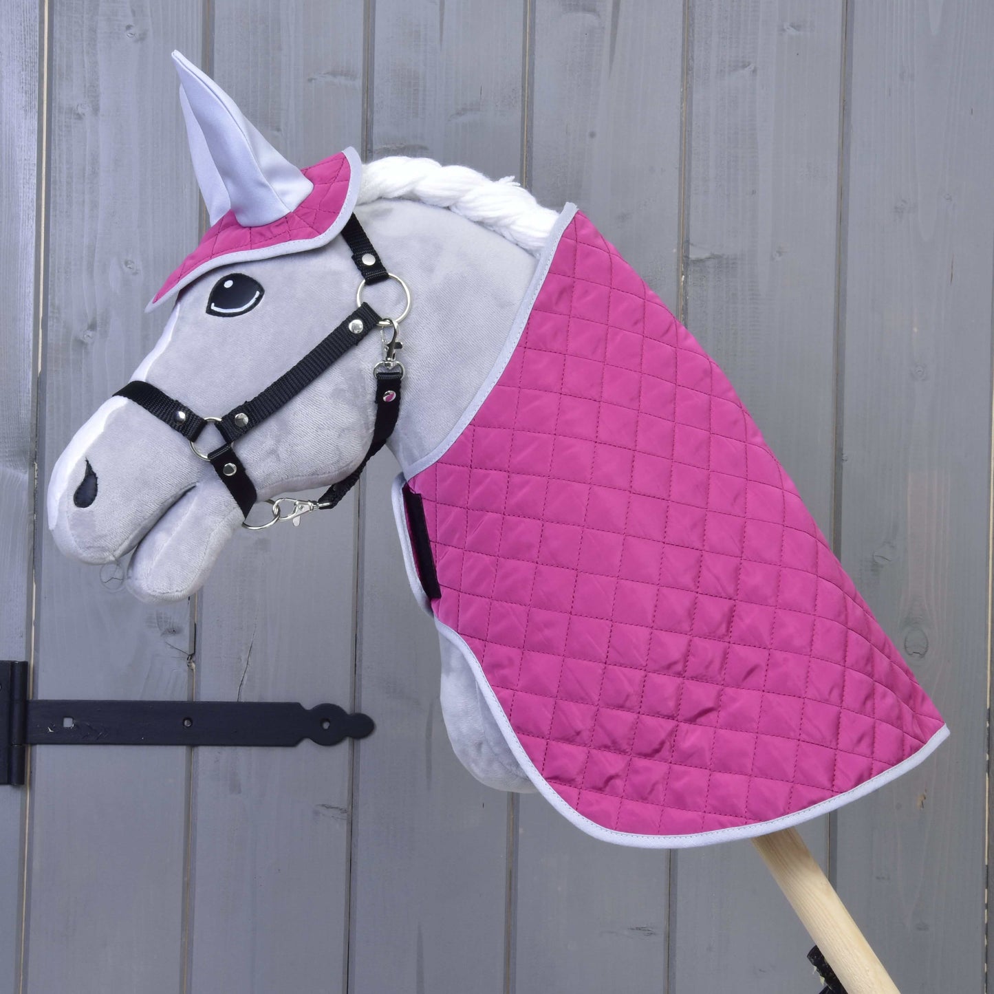 Das Hobby Horse LarDen Set Hot Pink Silver zeigt ein plüschiges Hobby Horse mit grauem Gesicht, schwarzem Zaumzeug, weißer Mähne, gestepptem Deckenset in Hot Pink und Weiß sowie passender Fliegenhaube, alles an einem Holzstab vor einer Holzwand.