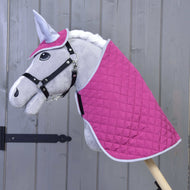 Das Hobby Horse LarDen Set Hot Pink Silver zeigt ein plüschiges Hobby Horse mit grauem Gesicht, schwarzem Zaumzeug, weißer Mähne, gestepptem Deckenset in Hot Pink und Weiß sowie passender Fliegenhaube, alles an einem Holzstab vor einer Holzwand.