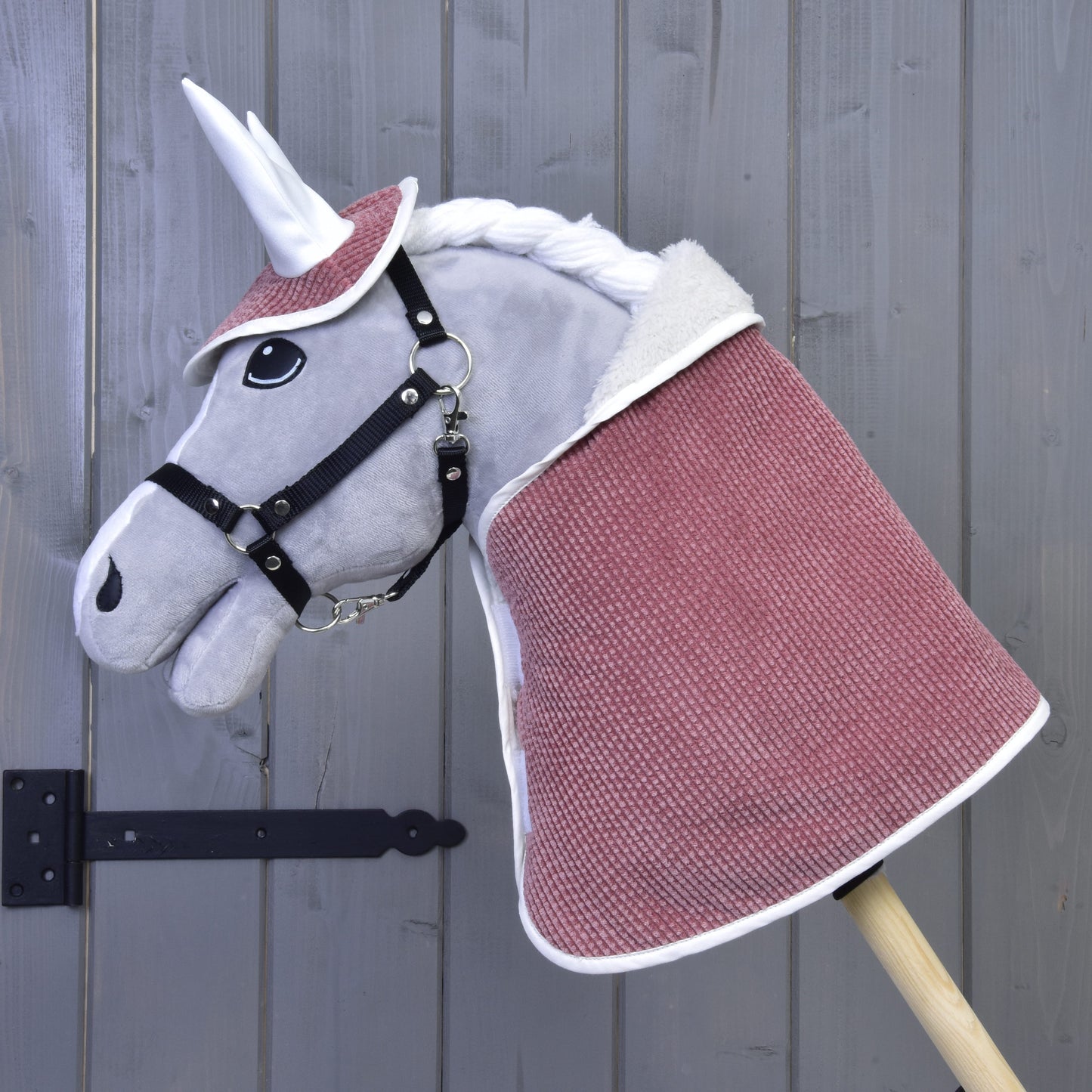 The Hobby Horse LarDen Set Manchester Tea Rose features a plush unicorn with a white horn, black bridle, pinkish-red covering, ear bonnet, and white mane on a wooden stick, displayed before a gray wooden wall.