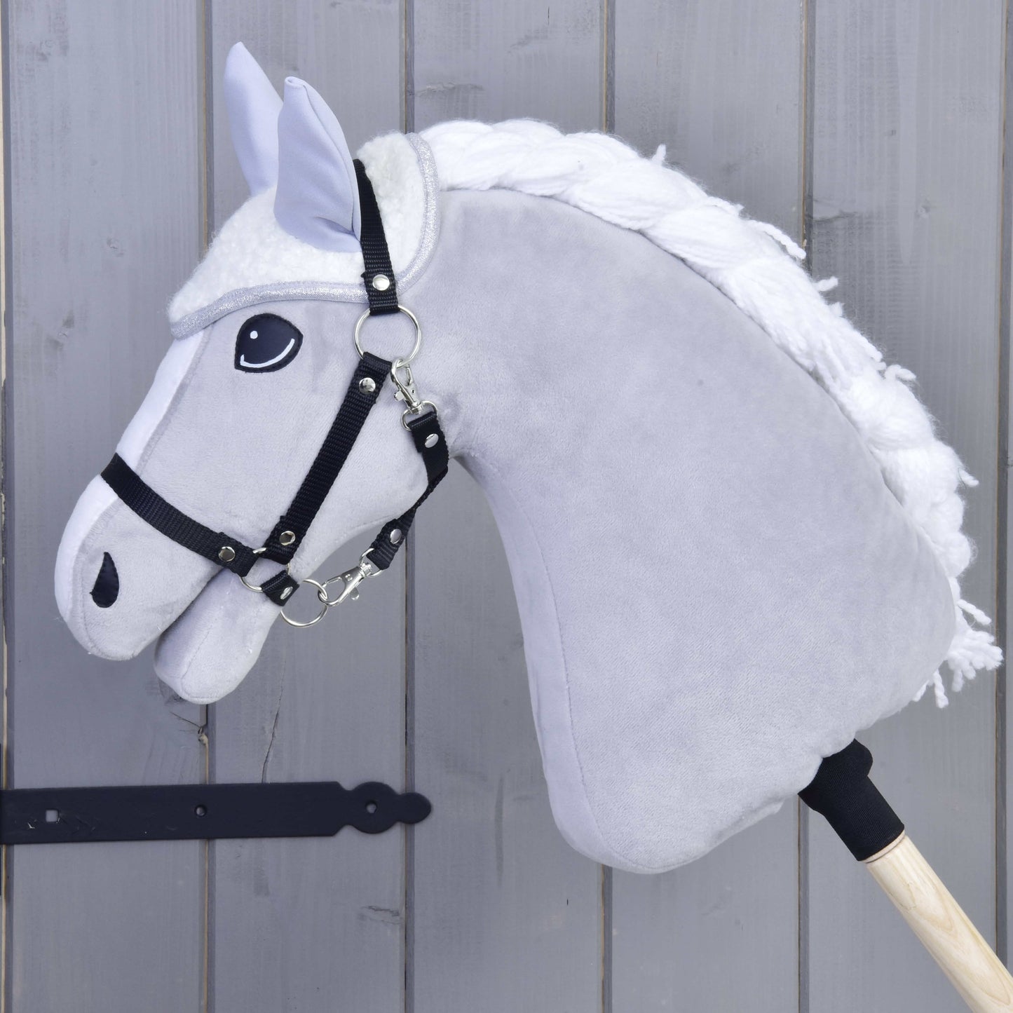 Nahaufnahme von Hobby Horse LarDen Set Snowflake mit grauem Körper, weißer Mähne, schwarzem Zaumzeug und Zügeln sowie einer Fliegenhaube auf einem Holzstab vor grauem Holzhintergrund.