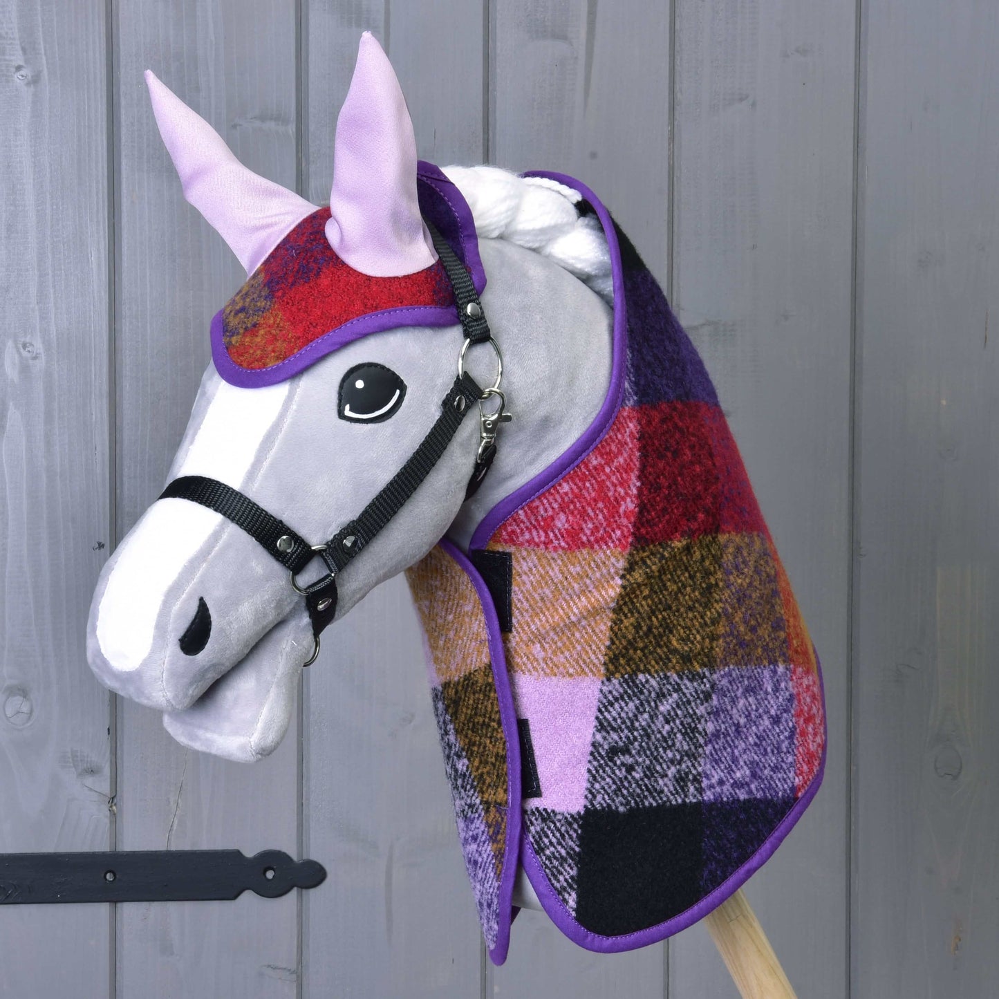 Set kariert Herbst 2 von Hobby Horse LarDen mit einem grauen Hobby Horse mit großen schwarzen Augen, ausgestattet mit einer karierten Decke in Rot, Lila, Gelb und Schwarz und passender Fliegenhaube auf einem Holzstab vor grauer Wand.