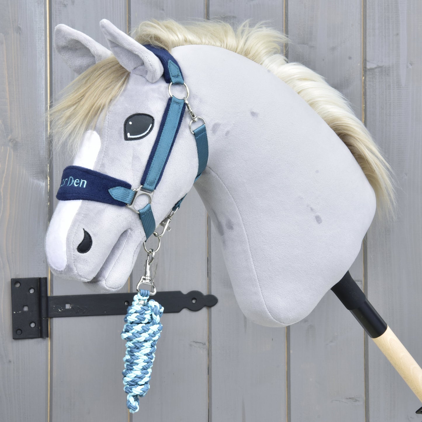 Set LarDen Exclusive Blue von Hobby Horse LarDen mit grauem Hobby Horse mit blauem Halfter für Hobby Horse und gedrehtem Führstrick in Blau-Weiß, auf einem Holzstab befestigt und vor einem Holzzaun-Hintergrund gezeigt.