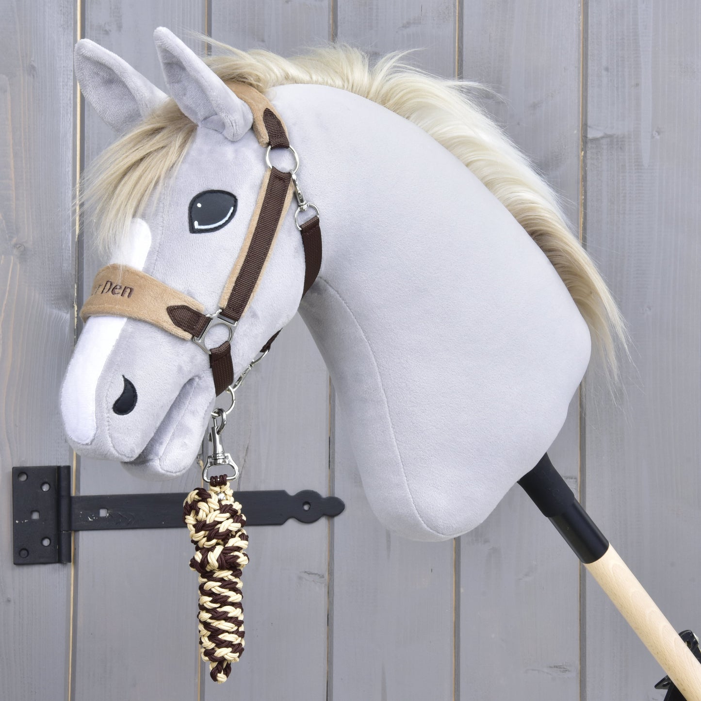 Set LarDen Exclusive Brown von Hobby Horse LarDen mit grauem Hobby Horse mit Zaumzeug für Hobby Horse, Fleece-Nasenriemen und creme-braunem geflochtenem Führstrick an einem Holzstab, fotografiert vor einer hellgrauen Holzwand.