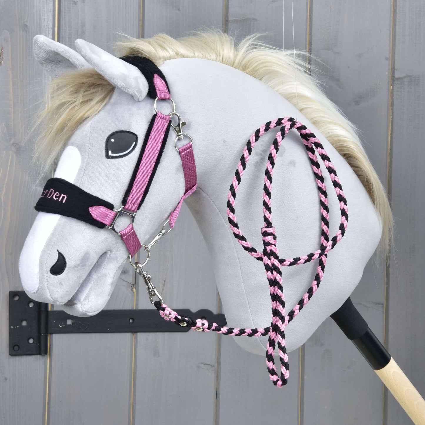 Set LarDen Exclusive Pink von Hobby Horse LarDen mit grauem Plüsch-Hobby Horse mit cremefarbener Mähne, Zaumzeug für Hobby Horse in Pink und Schwarz, passendem Fleece-Nasenriemen und Führstrick an einem schwarzen Stock, vor Holz-Hintergrund.