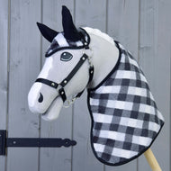 Das Hobby Horse LarDen Set Fleece Double-Sided zeigt ein plüschiges Hobby Horse mit schwarz-weiß karierter Hals- und Kopfabdeckung, schwarzem Zaumzeug, schwarzen Ohren und einem Fliegenhauben-Set, vor einem grauen Holzbackground.