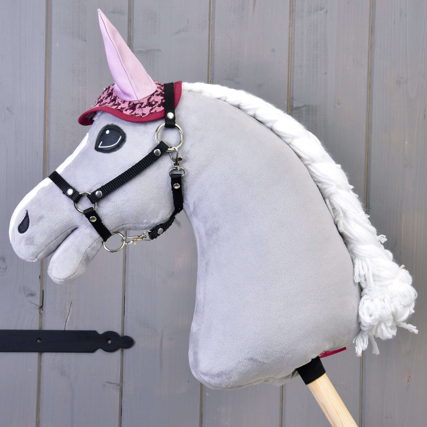 Ensemble festif Bordeaux de Hobby Horse LarDen sur un Hobby Horse gris en peluche avec crinière blanche, oreilles roses et bride rouge, portant une couverture élégante, monté sur un bâton en bois devant un fond en bois gris.