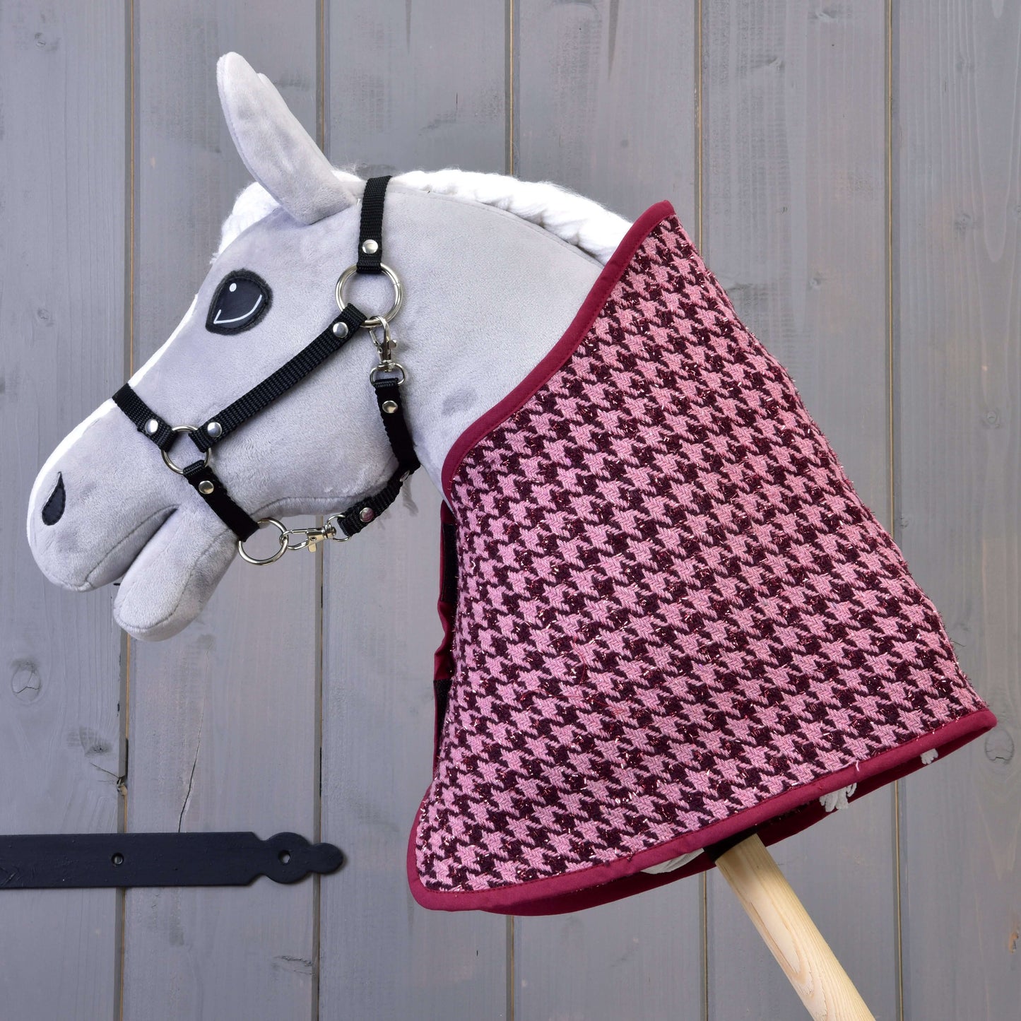 Ensemble festif Bordeaux de Hobby Horse LarDen, Hobby Horse gris en peluche avec bride noire et couverture pied-de-poule bordeaux et rose, monté sur un bâton en bois comme décoration charmante devant un fond en bois gris.