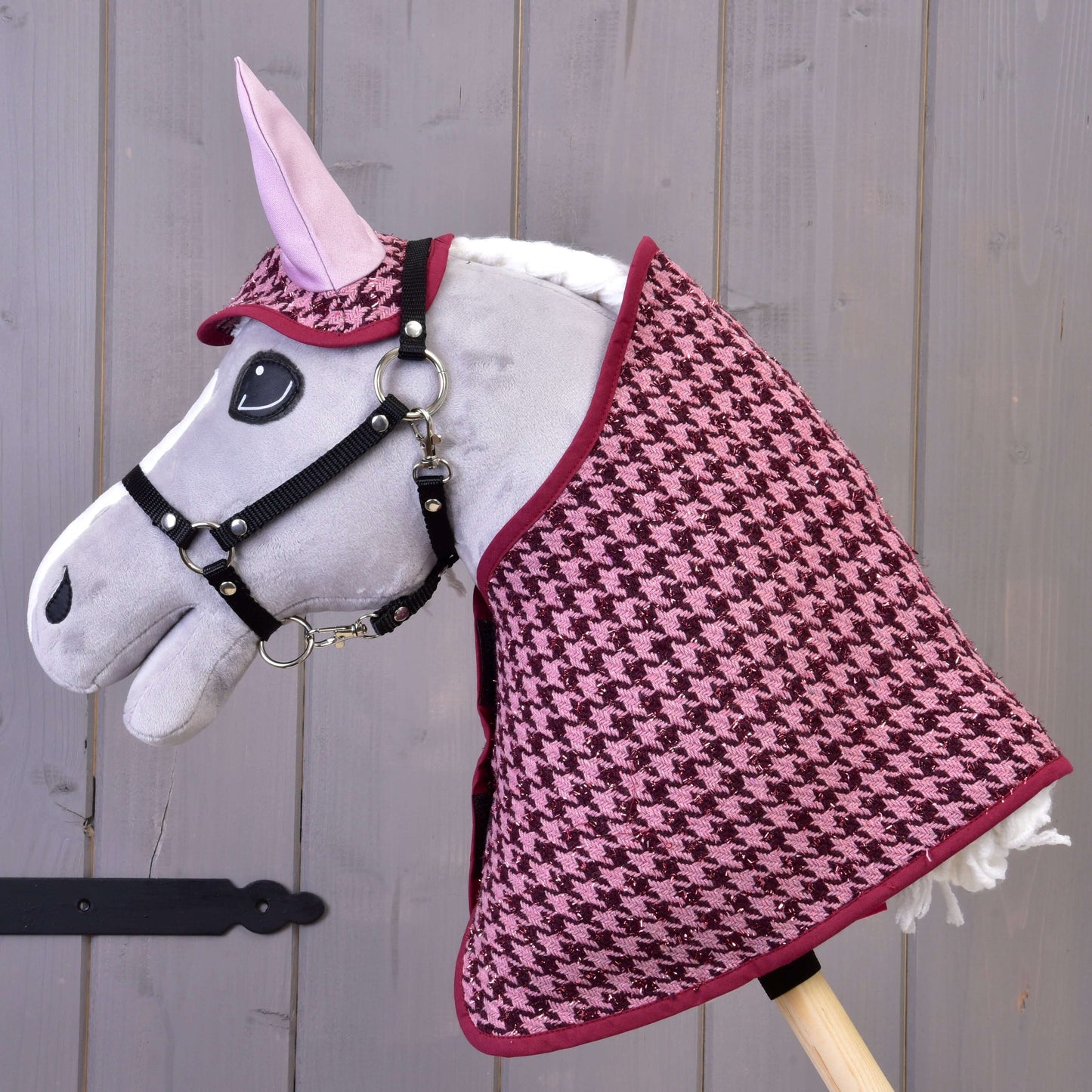 Cheval bâton gris de Hobby Horse LarDen portant l'ensemble couverture et bonnet d'oreilles Ensemble festif Bordeaux à motif pied-de-poule rose et noir, parfait comme décoration de cheval, posé devant un mur en bois gris.