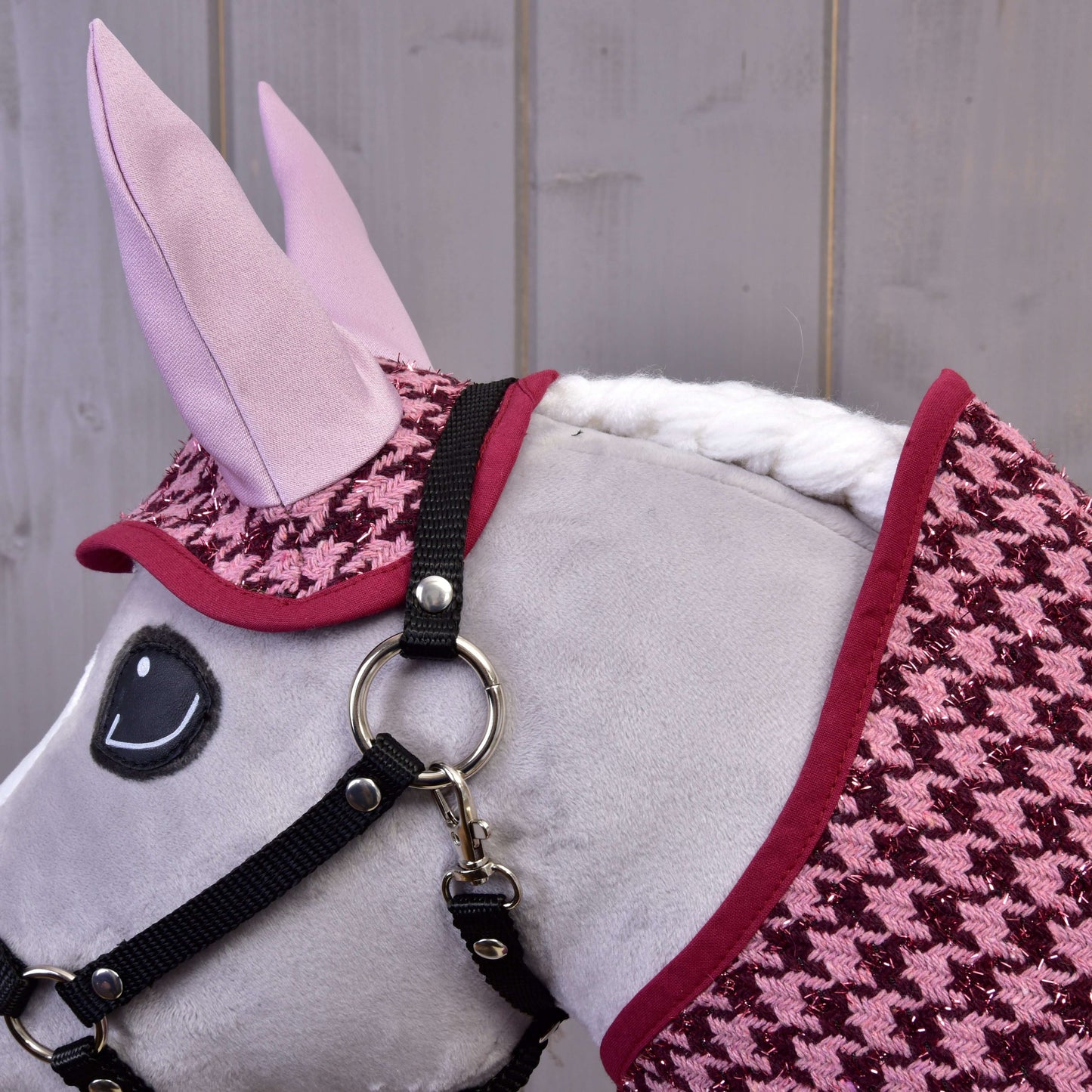 Gros plan d'un cheval jouet en peluche portant une bride noire et l'ensemble couverture et bonnet d'oreilles Ensemble festif Bordeaux de Hobby Horse LarDen, devant un mur en bois, créant une charmante décoration de cheval.