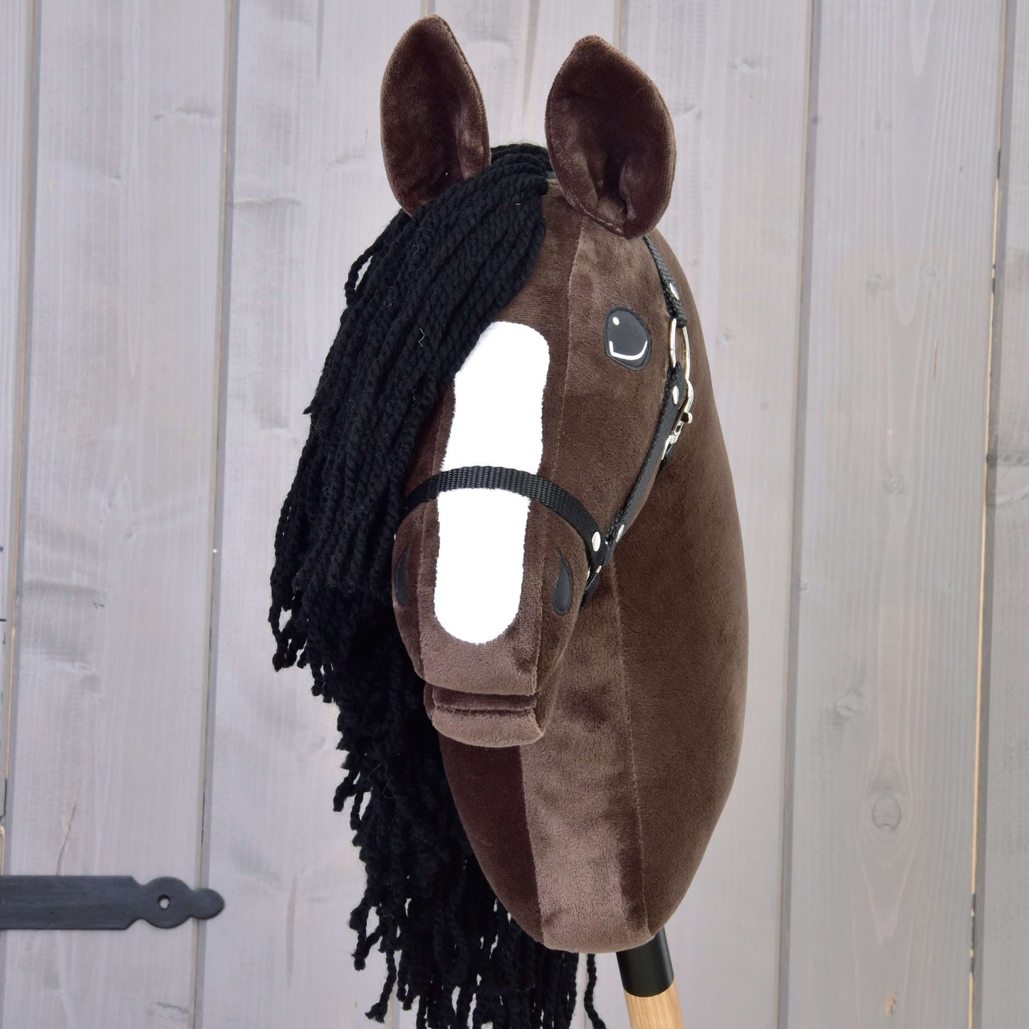 Hobby Horse Sisi avec licol noir de Hobby Horse LarDen, avec visage brun foncé, marque blanche sur le museau, crinière en fil noir et bride, capturant l’élégance d’un cheval de sport hanovrien sur fond en bois clair.