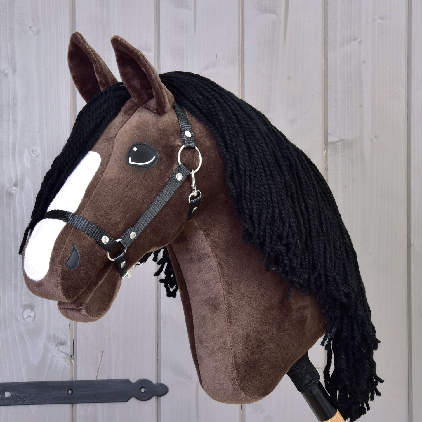 Hobby Horse Sisi avec licol noir de Hobby Horse LarDen, avec fourrure brun foncé, liste blanche sur le visage, crinière en fil noir et ressemblant à un jeune cheval de sport hanovrien, présenté sur fond de panneaux en bois.