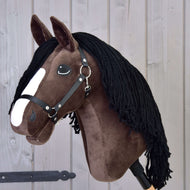 Hobby Horse Sisi mit Schwarzem Halfter von Hobby Horse LarDen mit dunkelbraunem Fell, weißem Gesichtsstreifen, schwarzer Garnmähne und im Stil eines jungen Hannoveraner-Sportpferds vor Holzpaneelen.