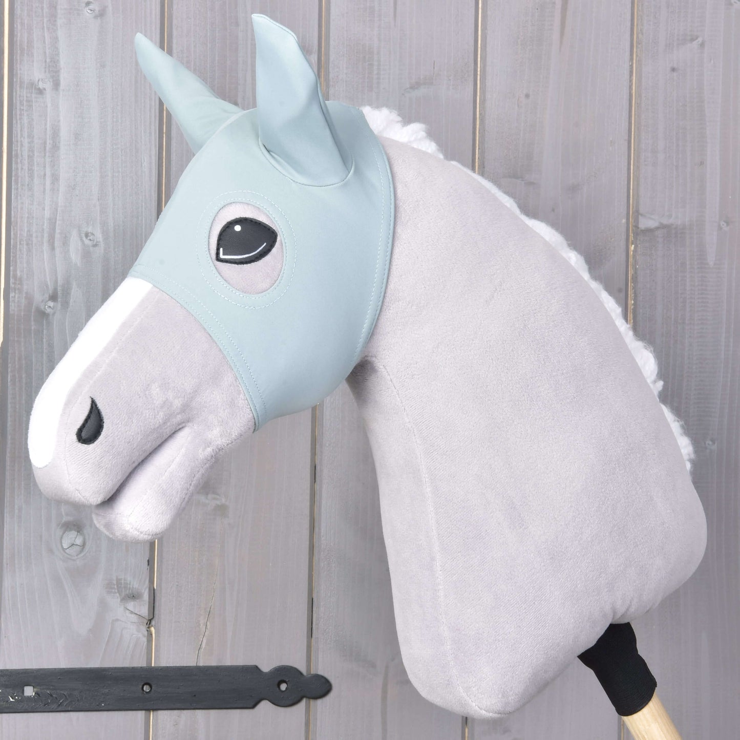 Das Hobby Horse LarDen „Maske mit Ohren Spiky“ zeigt ein graues Plüschpferd mit hellblauem Zaumzeug, weißer Mähne und Schutzmaske auf einem Holzstab, präsentiert vor einem Hintergrund aus hellen Holzplanken.