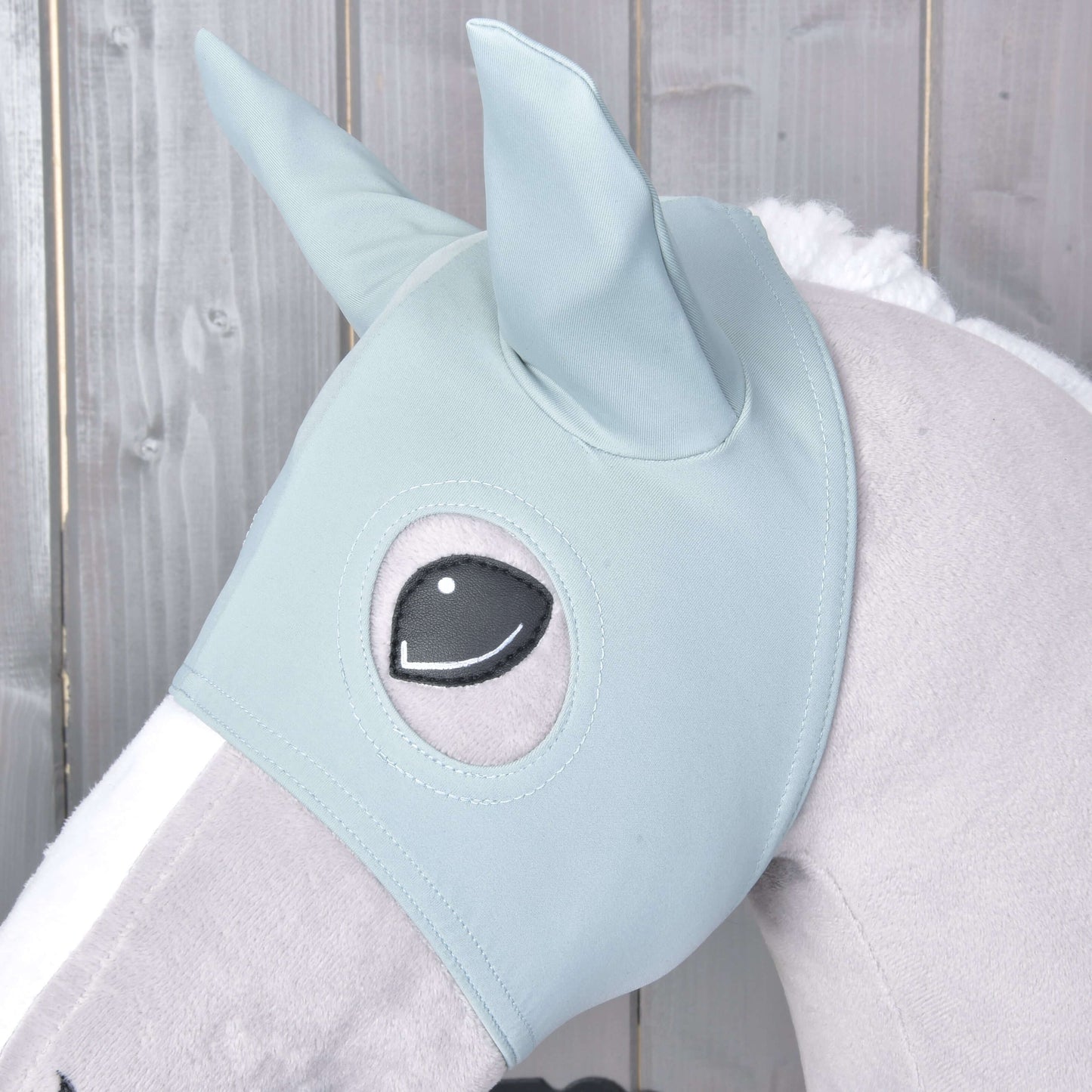Nahaufnahme der Maske mit Ohren Spiky von Hobby Horse LarDen in Hellblau mit Ohrabdeckungen und einer Augenöffnung, gezeigt auf einem Plüschtier vor einem Holzhintergrund.