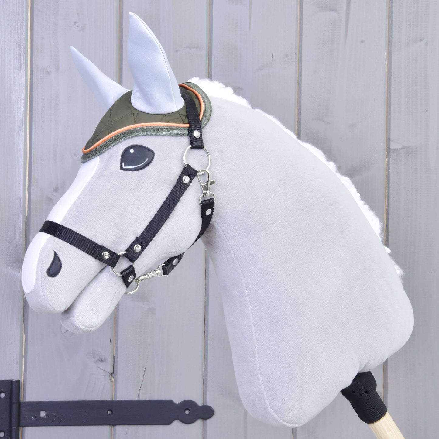 Hobby Horse en peluche de Hobby Horse LarDen sur bâton avec crinière blanche, visage gris, bride noire et œil brodé, portant le Bonnet d'oreilles Sport, un bonnet d’oreilles vert à bordure orange, sur fond en bois clair.