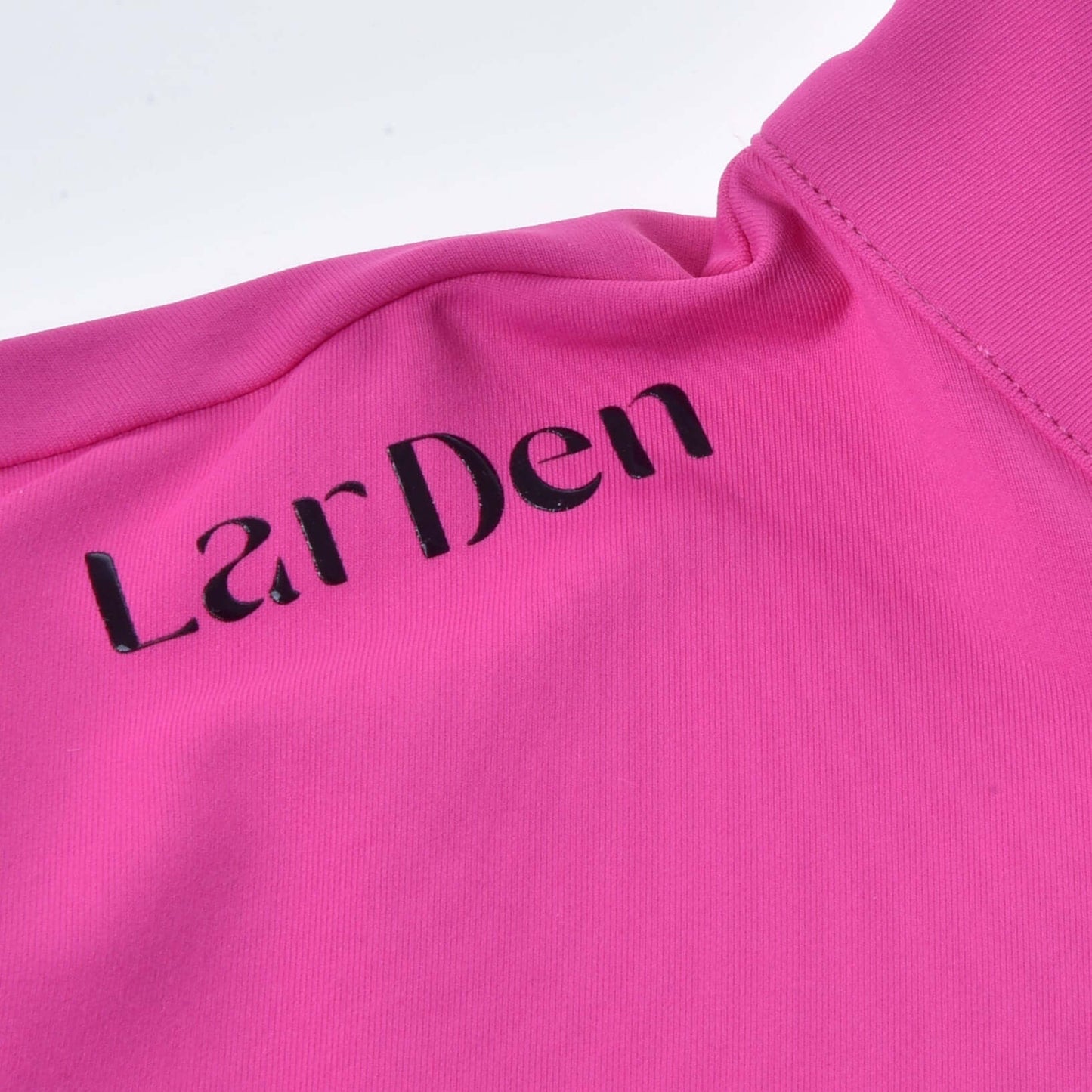 Nahaufnahme des T-Shirt LarDen Pink von Hobby Horse LarDen mit kurzen Ärmeln, glattem Spandex-Stoff, sichtbaren Nähten und Struktur sowie schwarzem „LarDen“-Aufdruck auf dem leuchtend pinken Material.