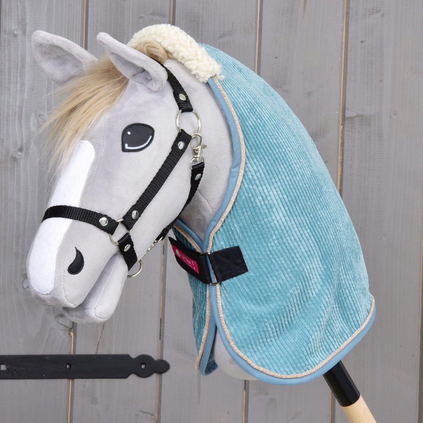 Ein Plüsch-Hobby Horse von Hobby Horse LarDen mit hellgrauem Gesicht, schwarzem Zaumzeug und weißer Schnauze trägt die Stable Blanket Fern aus hellblauem PES-Material und ist vor Holz-Hintergrund positioniert.