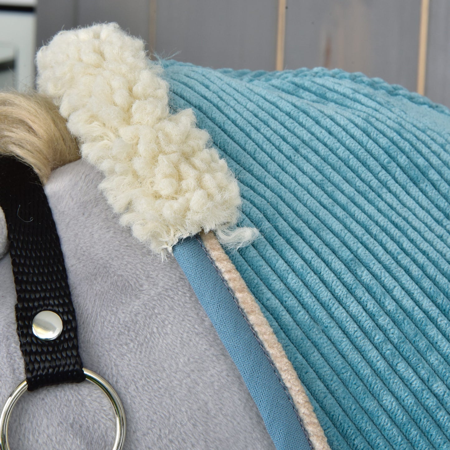 Nahaufnahme eines hellgrauen Plüschpferds mit der blauen Stable Blanket Fern von Hobby Horse LarDen mit cremefarbenem Fleecebesatz und schwarzem Riemen mit silbernem Ring. Im Hintergrund sind helle Holzpaneele sichtbar.