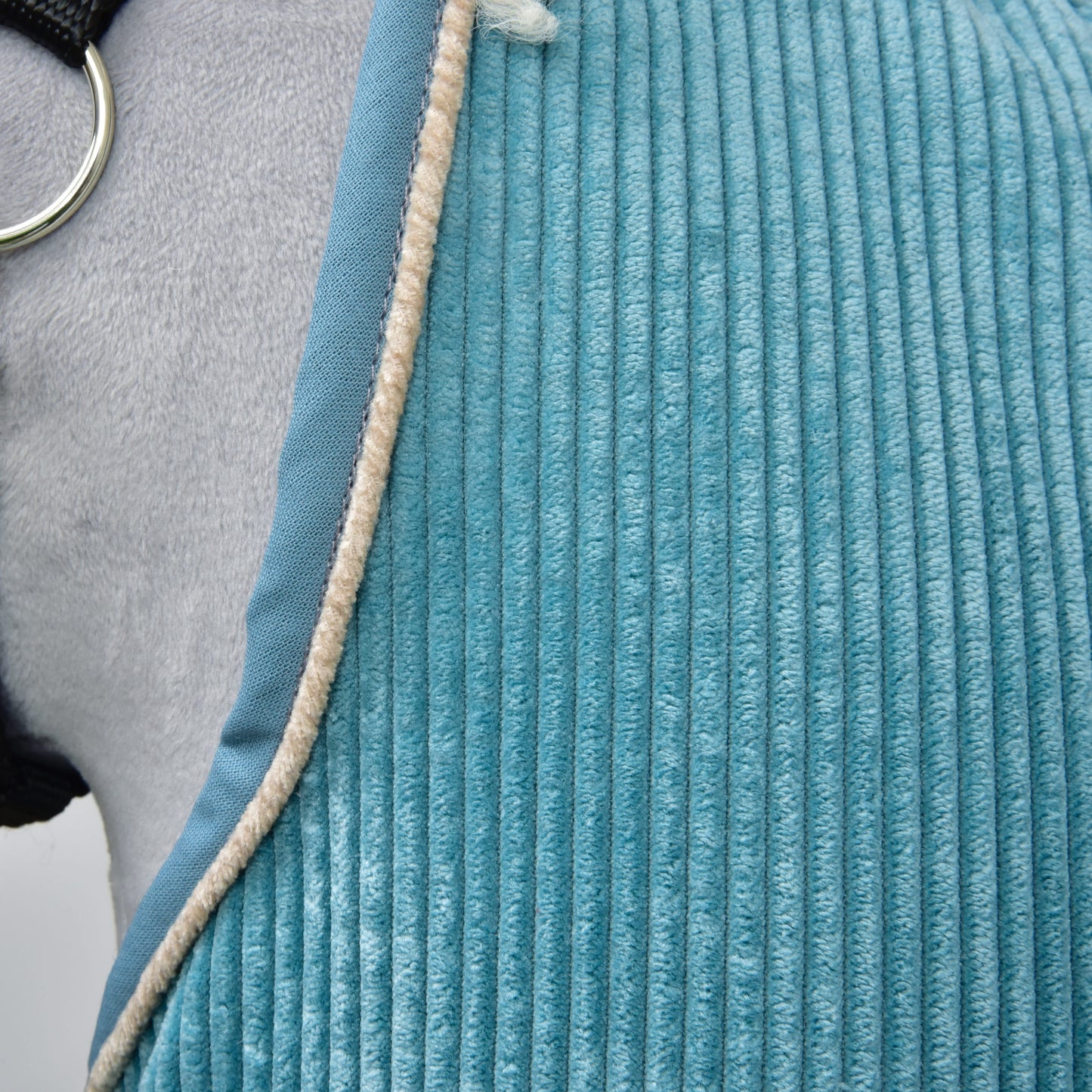 Nahaufnahme der strukturierten blauen Stable Blanket Fern von Hobby Horse LarDen, über ein graues Pferd gelegt, mit beigefarbener flauschiger Kante und sichtbarem Metallring des Pferdegeschirrs.