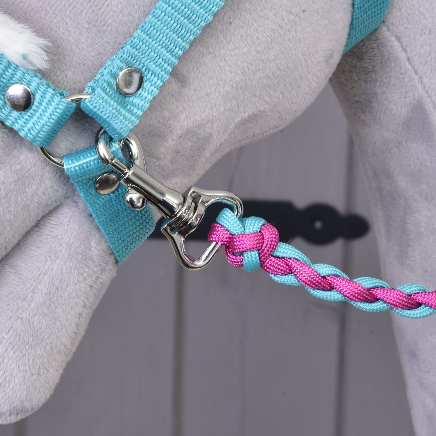 Gros plan sur la longe de l’Ensemble Combinaison Parfaite Turquoise de Hobby Horse LarDen, tressée en bleu et rose et fixée à un harnais bleu clair sur un animal en peluche gris, idéale comme accessoire pour Hobby Horse. Arrière-plan flou.
