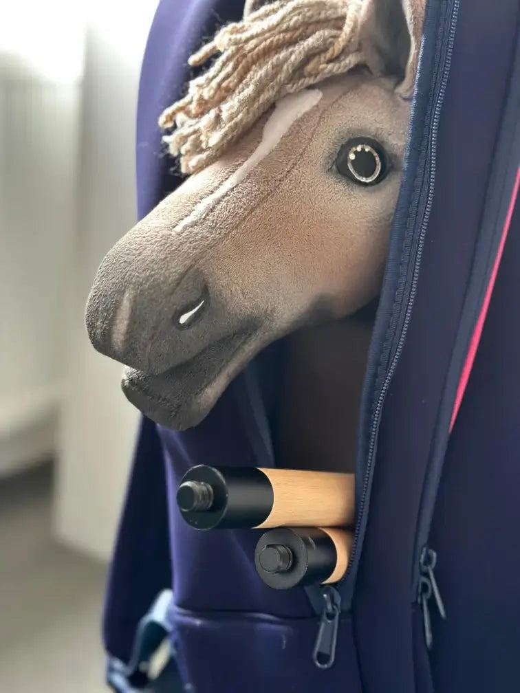 Ein Plüsch-Hobby Horse mit hellbrauner Mähne und weißer Blesse schaut aus einer teilweise geöffneten dunkelblauen Tasche, in der auch zwei Holzstiele sichtbar sind – perfekt für Hobby Horsing-Abenteuer.