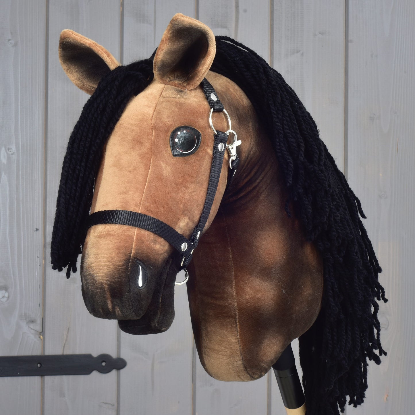 The Hobby Horse LarDen "Hobby Horse Stormy with Black Halter," featuring a plush brown body and a long black yarn mane, is posed in front of a light gray wooden wall.