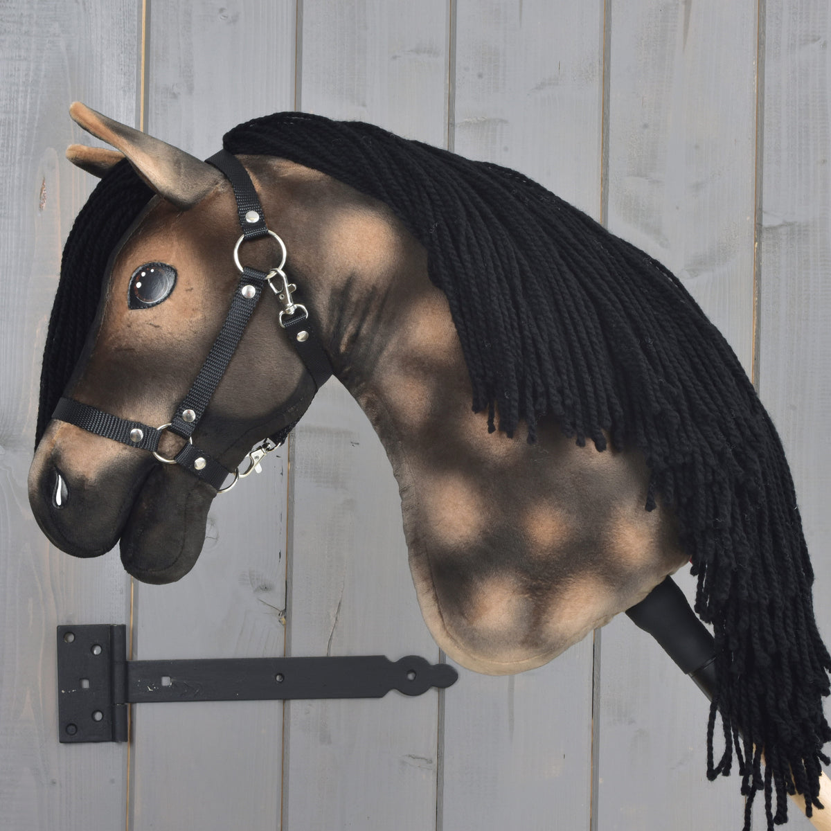 A realistic Hobby Horse Stormy with Black Halter from Hobby Horse LarDen, inspired by a Hanoverian Stallion and featuring a black yarn mane, is mounted on a gray wooden wall with a black bracket.